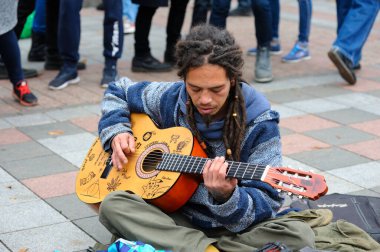 Sokak ortasında oturan, akustik gitar çalan, arka planda yürüyen yaya bacaklı sokak müzisyeni rastaman. 26 Ekim 2019. Kyiv, Ukrayna