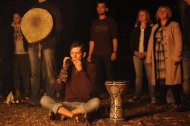 Young man, the follower of Ukrainian pagan cult, playing darbuka drum, people standing around, light from the burning fire set. November 11, 2019. Kiev, Ukraine clipart