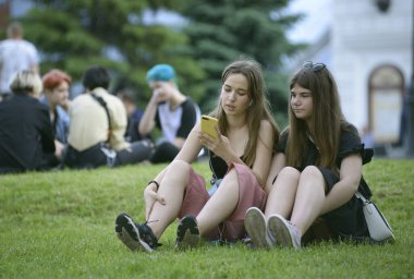 Şehir parkında çimenlerde oturan ve cep telefonlarına bakan kızlar. 25 Mayıs 2019. Kyiv, Ukrayna