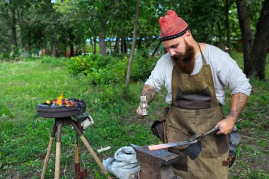 Antik Slav demirci gibi giyinmiş demirci adam yanan bir demircide metal dövüyor, örsle çalışıyor. 2 Ağustos 2018. Murometz Parkı, Kyiv, Ukrayna