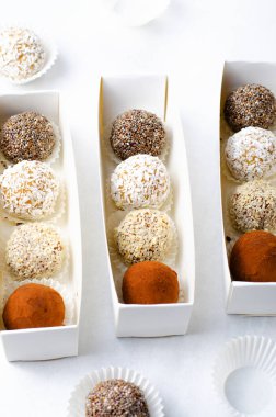 Çeşitli vejetaryen tatlılar, tohumlu lezzetli şeker topları, kurutulmuş meyve ve kakao tozu, Chia 'lı sağlıklı şeker, susam tohumları, Hindistan cevizi ve mantarlar parlak arkaplanda karton kutularda.