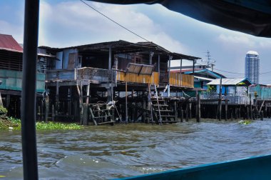 Stilt houses on the Chao Phraya River in Bangkok, Thailand. Contrast of traditional dwellings and a modern skyscraper.