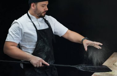 Önlüklü ve bereli bir şef kürekle un serpiştirir, siyah arka planda yakın plan.