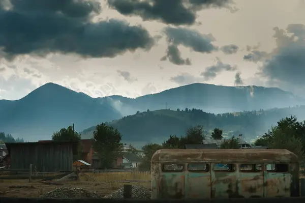 stock image Carpathian mountain landscape, an abandoned yard, a rusty wagon, in the background silhouettes of mountains illuminated by the rays of the sun from a dark sky