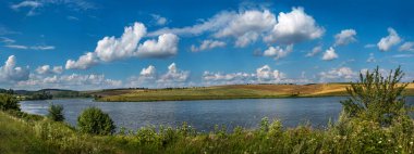 Rezervuar boyunca panoramik bir manzara tepedeki tarlalara ve mavi gökyüzünün altında bulutlu tahıl ekinlerine. Yazın göl kenarında sakin bir kırsal.
