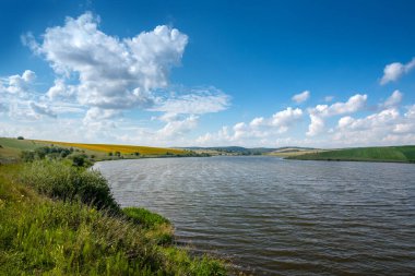 Yazın göl ya da nehir kıyısında sakin bir kırsal. Rezervuar boyunca uzanan panoramik bir manzara bulutlu açık mavi gökyüzünün altındaki tepelik alanlara doğru.