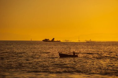 Bir tekne silueti ve bir itfaiye uçağı gün batımında deniz suyuna iniyor. Yangını söndürmek için depoyu suyla dolduruyorlar.