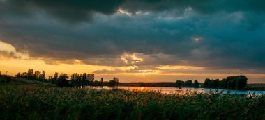 Gölün ya da nehrin üzerinde gün batımının manzarası. Gölün üzerindeki kara bulutlar batan güneşin sıcacık ışığıyla suya yansıyan bir tezat oluşturuyor..