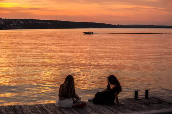Ternopil, Ukrayna - 30 Ağustos 2020: Ternopil 'deki bir gölün tahta iskelesinde sıcak bir akşamda iki kız arkadaşın silueti. Turuncu bir gökyüzünün arka planında, sakin sularda bir tekne, huzurlu bir atmosfer..