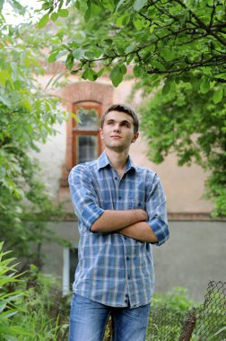 Kareli gömlekli genç bir adamın portresi, bahçedeki eski bir evin arka planında düşünceli bir şekilde duruyor..