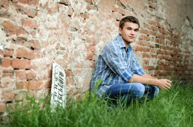 Kareli gömlekli ve kotlu şık bir genç adam. Tuğla duvarın yanındaki çimlerin üzerinde düşünceli bir şekilde oturuyor.