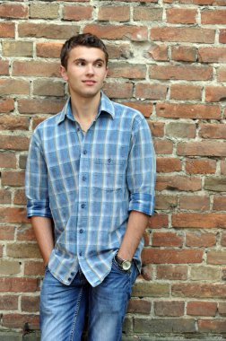 Kareli gömlekli, kotlu şık bir genç adam elleri ceplerinde, sırtı eski tuğla bir duvara dönük duruyor.