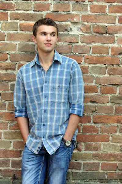 Kareli gömlekli, kotlu şık bir genç adam elleri ceplerinde, sırtı eski tuğla bir duvara dönük duruyor.