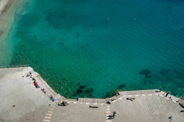 Rıhtımın üst manzarası kristal berrak turkuaz su ve deniz kenarında dinlenen insanlar. Sakin atmosfer, temiz sahil ve parlak güneş ışığı mükemmel bir yaz havası yaratır..