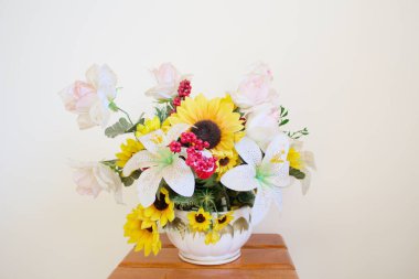 Beyaz bir duvarın önünde sarı ve beyaz plastik çiçekli bir vazonun yatay, durgun bir yaşam stüdyosu çekimi. Bahar zamanı ayçiçeği düzenleme.