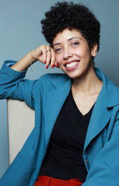 Başarılı bir iş kadını koltukta rahatça oturup kameraya gülümsüyor. Kısa kıvırcık saçlı Afro-Amerikan kadın model mavi ceket ve siyah gömlek giyiyor..