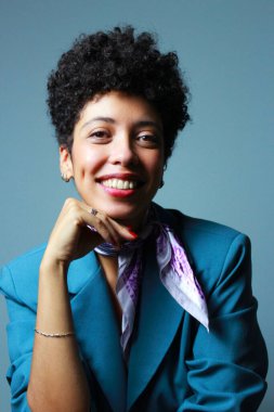 Başarılı bir iş kadını, elinde çenesiyle kameraya parlak bir şekilde gülümsüyor. Kısa kıvırcık saçlı Afro-Amerikan kadın model mavi ceket ve leylak atkı giyiyor..