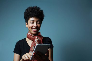 Afro-Amerikalı neşeli bir kadın tabletten e-kitap okuyor. Ön mavi arka planda kısa saçlı bir kadın model ve fotokopi alanı var. Edebiyat, araştırma, web araması, online aktivite reklamları için elverişli.