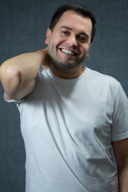 Beyaz tişörtlü çekici bir adam, eli boynunda gülümsüyor. Mutluluk, flört, sadelik, erkeklerin refahı için idealdir. Gri arka planlı dikey stüdyo portresi.