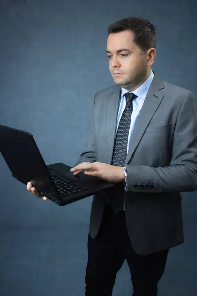 Gri takım elbiseli, ayakta duran ve dizüstü bilgisayarını kullanan odaklanmış bir iş adamı. Çevrimiçi iş, ofis işi, teknoloji ve şirket yönetimi için ideal. Mavi arkaplanda kopya alanı olan stüdyo portresi.