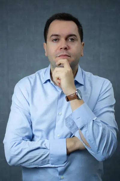 Eli çenesinde, mavi gömleği ve düğmesiz kelepçeleri olan ciddi bir iş adamı. Kurumsal yaşam tarzı, ofis işi, menajer ve iş teması için ideal. Mavi arkaplan ile stüdyo portresi.