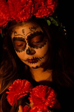 Koyu arkaplanda Catrina gibi giyinmiş bir kadın elinde tuttuğu kırmızı çiçeklere bakıyor.