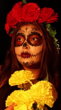 Catrina gibi giyinmiş bir kadın elinde bir buket sarı çiçek tutuyor.