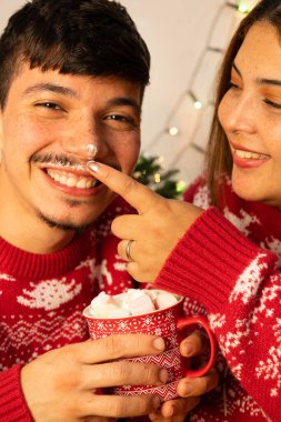 Neşeli çift Noel 'de eğlenirken marşmelovla sıcak çikolata içip arka planda ağaç ve Noel ışıkları olan kazaklar giyiyorlar.