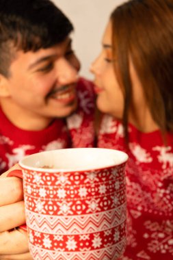 Odaklanamayan neşeli çift Noel arifesinde ellerinde bir fincan çikolata tutuyorlar ve Noel kazakları giyiyorlar.