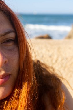 Düşünceli ve sorgulayıcı bir ifadeyle çilli genç kızıl saçlı kadın güzel beyaz bir kumsalda oturuyor ve arka planda muhteşem bir deniz ve yanında da güzel açık mavi bir gökyüzü.