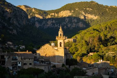 Valldemossa kasabasının sabahki görüntüsü. Kilisenin ve antik yapıların bulunduğu yerde, yapraklı ağaçlarla dolu etkileyici dağların arkasında.