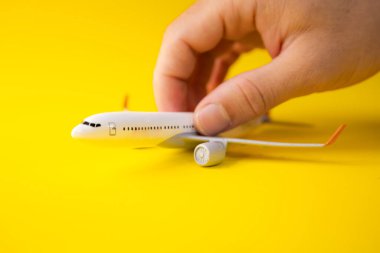 Sarı arka planda bir adamın parmakları tarafından tutulan minyatür bir uçağın yan görüntüsü, ulaşım ve uçuşlar kavramı