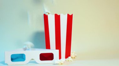 Patlamış mısır yavaş çekimde karton kovaya düşüyor ve yanında mavi ve kırmızı 3D gözlükler, sinema ve eğlence konsepti var.