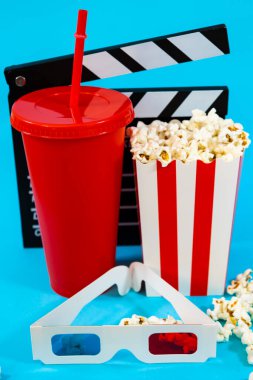 Kırmızı soda bardağının arkasındaki film sepetinin dikey görüntüsü, bir kova patlamış mısır ve mavi arka plandaki 3D gözlükler, sinema eğlencesi konsepti