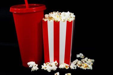Beyaz ve kırmızı çizgili bir karton kovanın yatay görüntüsü. Patlamış mısır ve bir bardak kırmızı soda ile siyah arka plan, sinema, yemek ve eğlence konsepti.