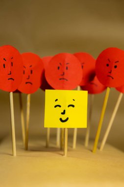 Vertical image of yellow paper with smiley face surrounded by angry faces, concept of social rejection and discrimination clipart