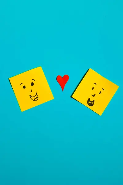 Mavi arka plan, arkadaşlık ve sendika kavramı üzerine çizilmiş gülen yüzlü ve kırmızı kalpli sarı kare kağıtların dikey görüntüsü.