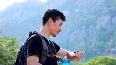 Maceracı genç adam saatine bakar ve güneşli bir günde dağlarla çevrili yürüyüşüne devam eder.