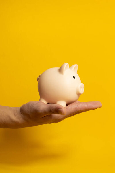 Vertical image of a male hand holding a piggy bank on a yellow isolated background, economy and finance concept