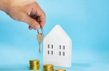 Erkek el, üzerinde altın ve parmaklar olan bir anahtar taşıyor ve yanında mavi arka planda beyaz ahşap bir ev, ev satın almak için tasarruf kavramı.