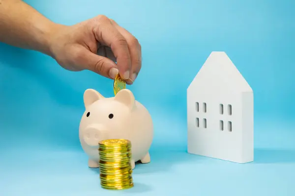 Erkek el, arkasında odağından beyaz ahşap bir ev olan altın paraların yanına bir kumbara koyuyor. mavi arkaplan, ev tasarrufu kavramı