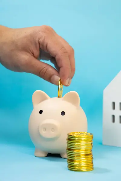 Erkeğin dikey görüntüsü, arkasında odağını kaybetmiş beyaz ahşap bir ev olan altın paraların yanındaki kumbaraya bozuk para koyması. mavi arkaplan, ev tasarrufu kavramı