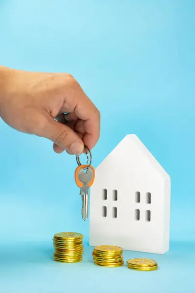 Dikey görüntü, parmakları olan bir erkek eli etrafında altın ve anahtar ve yanında mavi arka planda beyaz ahşap bir ev, ev satın almak için tasarruf kavramı.