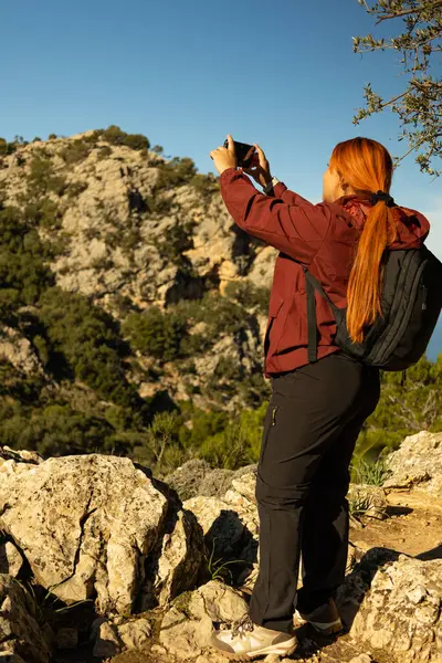 Doğanın tadını çıkaran bir dağın tepesinde cep telefonuyla fotoğraf çeken bir kadının dikey görüntüsü.