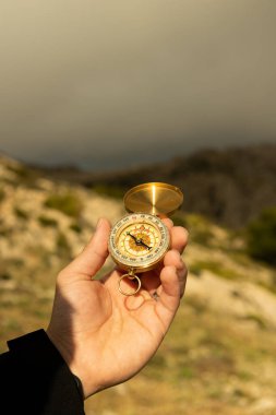 Erkek elinin dağda pusula tutarken dikey görüntüsü. Arka planda gri bulutlar, doğada yön bulma.