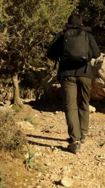 Güneşli bir günde dağlarda kayalık bir yolda yürüyen erkek yürüyüşçünün dikey videosu.