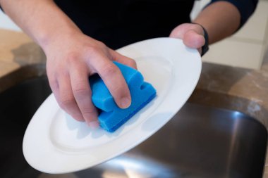 Mutfak lavabosunda mavi bir süngerle bulaşık yıkayan erkeklerin ellerini kapatın.