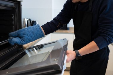 Adam evde yemek tepsisi ve mavi fırın eldiveniyle yemek pişiriyor.