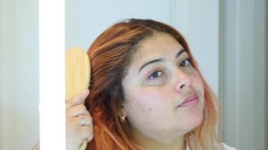Aynanın önünde kızıl saçlı, çilli bir kadının saçını ahşap bir çevre dostu tarağıyla taramasının videosunu kapat.