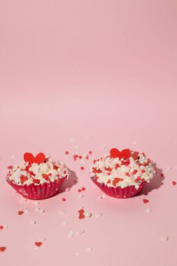 Vertical image of two decorated Valentine's Day cupcakes with sugar hearts scattered on a pink isolated background with copy space. Love and affection concept clipart